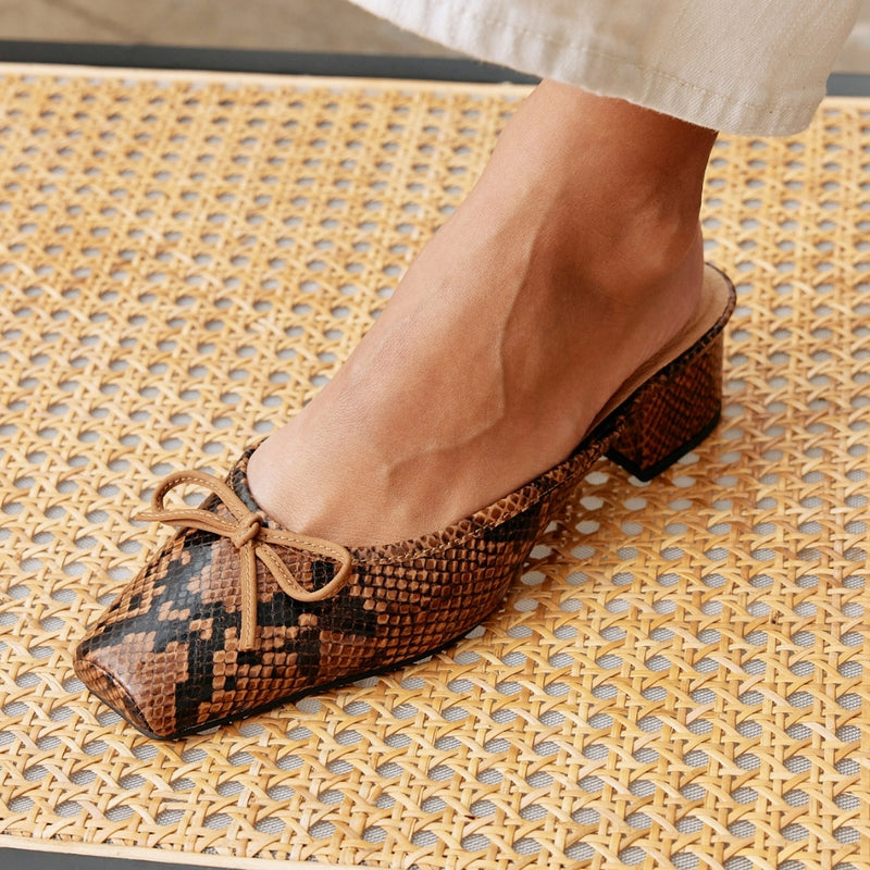 Close-up shot of a woman’s foot wearing a brown snake print mule heel with a square toe and bow detail. Perfect for adding a trendy touch to any outfit.