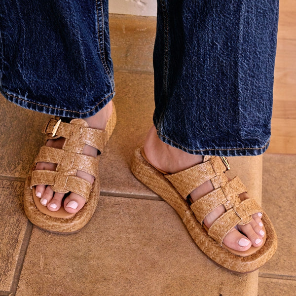 Tan raffia platform slide sandals with woven straps and an adjustable buckle detail. A breezy, laid-back style perfect for warm-weather wear.